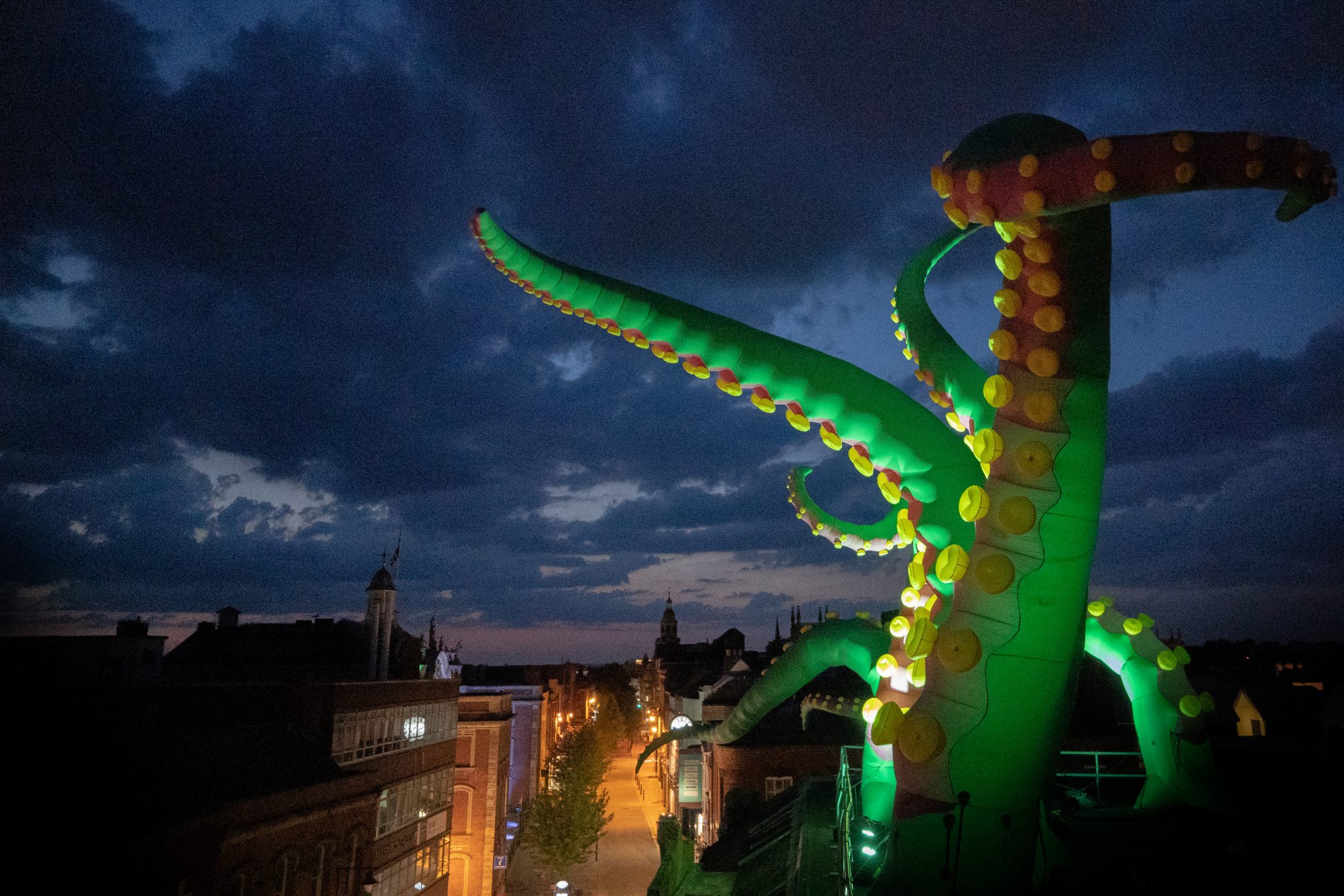 20210718-RAVPhotography-SameButDifferent-11524 Image Description: : Photo taken at night from the roof of a building where large green inflatable tentacles are lit up and are reaching out in different directions. Below you can see the high street shops lit by street lights.